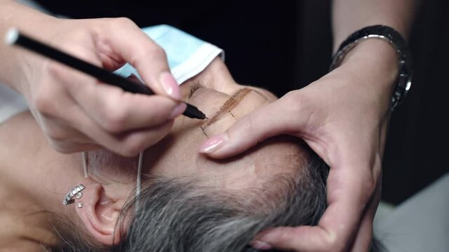 Female beautician making eyebrows correction with special make-up pigment pencil, cosmetology artist preparing middle-aged woman's face for microblading procedure in beauty salon. Facial contour