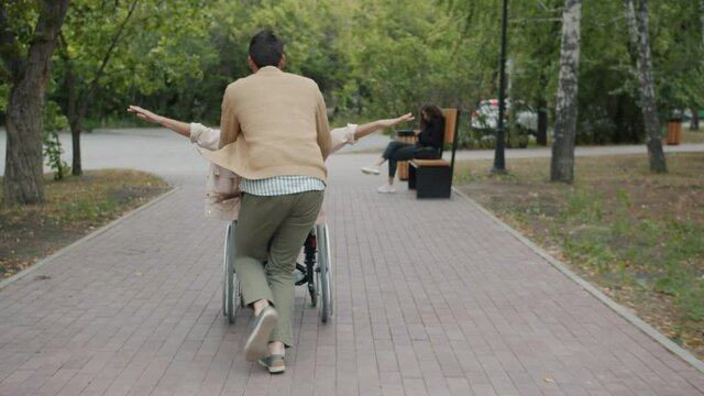 Caring husband is running and pushing wheelchair having fun with disabled wife outside in autumn park. Handicapped people and happy family concept.