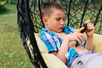 Boy in plaid shirt sitting in cocoon chair and playing with telephone. Smartphone games. Summer time