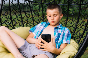 Boy in plaid shirt sitting in cocoon chair and playing with telephone. Smartphone games. Summer time