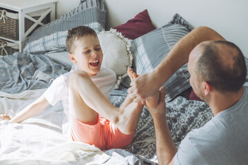 Father plays with his son 7-10 on bed. Dad tickles kids feet. Family, having fun - obrazy, fototapety, plakaty