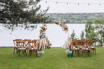 Wedding arch decorated with flowers, wooden chairs stand on green grass against the background of...