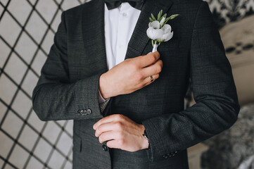 A man, a groom in a gray checkered suit straightens his boutonniere and bow-tie with his hand. Wedding portrait.