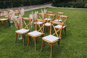 A row of wooden chairs with reed decorations stands on the green grass in the garden. Wedding photography.