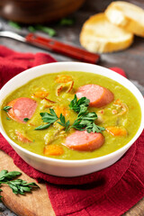 Green pea soup with carrots, potatoes, celery, sausages, pork and bacon in a white bowl on a dark wooden background. Snert is a Dutch pea soup.