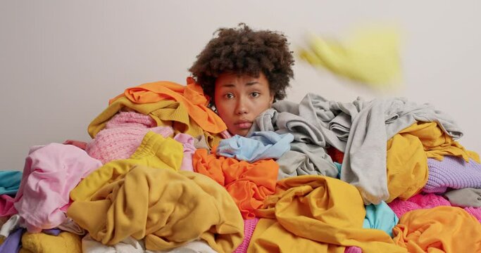 Unhappy curly haired Afro American woman with falling laundry on head poses around multicolored clothes isolated over white background. Busy housewife has much housework. Heap of dirty clothing