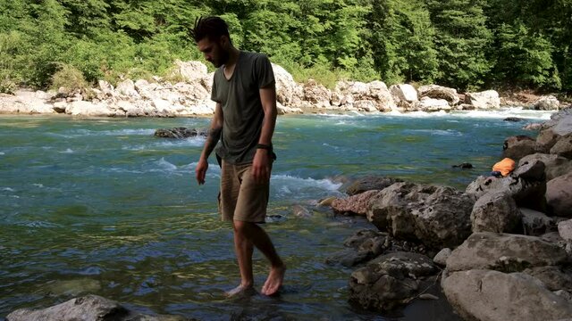Young handsome Caucasian man with beard and dreadlocks walks barefoot along bank of cold stormy mountain river and enjoys life. Human in shorts and Tshirt is wetting his feet in river. 4k footage.