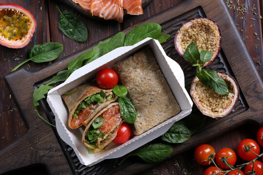 Food for delivery. Pancakes with with salmon, spinach and passion fruit dessert in a box on a gray wooden table. Rustic. Background image, copy space