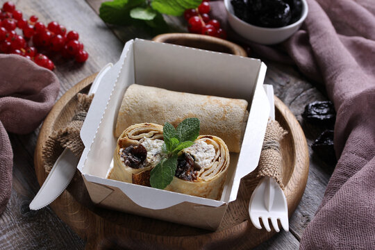 Food for delivery. Pancakes with cottage cheese and prunes and mint in a box on a gray wooden table. Rustic, red currant. Background image, copy space