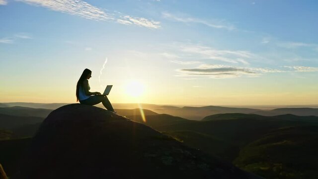 Aerial shots of a silhouette of a young freelance woman working on a laptop sitting on top of a rocky mountain at sunset.