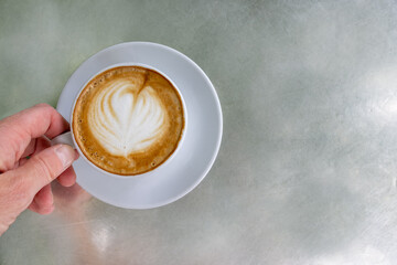 A woman's hand is holding a cup of cappuccino against a silver background with copy space 