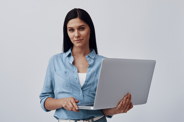 Attractive young woman using laptop and looking at camera while standing against grey background