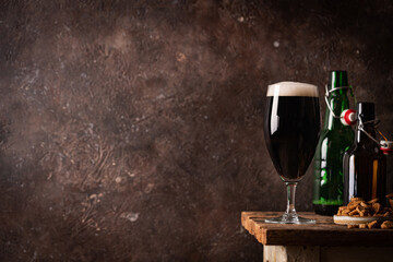 Glass of dark beer with foam head on dark wooden background with empty bottles and beer snacks