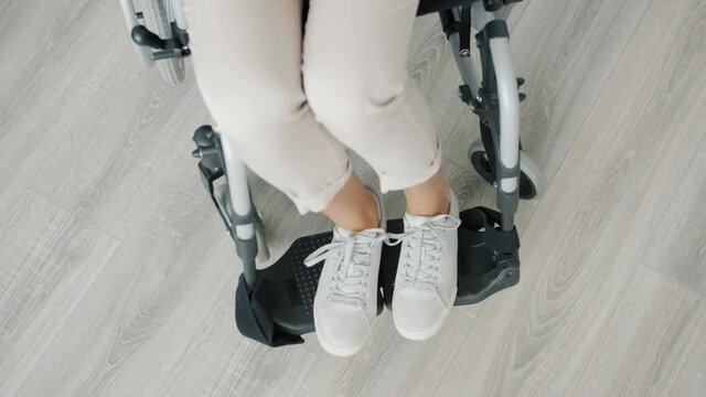 High angle view of female feet of paraplegic woman sitting in wheelchair indoors alone with wooden floor in background. People and disability concept.