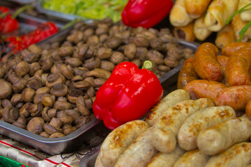 Small mushrooms lie on a large tray next to red bell peppers, various meat sausages and other foods. Delicious food cooked over an open fire, which is offered at a street food fair, event, festival.