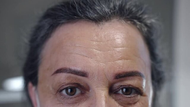Close-up portrait of happy middle-aged woman looking on camera in beauty salon after rejuvenate procedures, satisfied woman showing corrected permanent eyebrows tattoo. Natural asymmetry of eyebrows