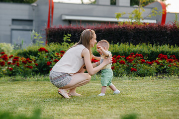 A young cute mother helps and teaches her little son to take his first steps during sunset in the park on the grass.