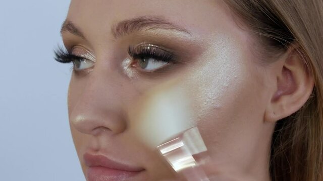 The highlighter is applied to the cheekbones of a beautiful young female model using a special thick brush. Professional make-up at the makeup artist's stylist in the studio