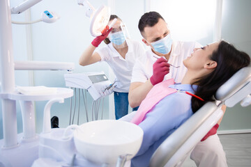 Male dentist in protective mask treat the teeth of a young patient. Woman sitting in a dental chair. A young assistant points a lamp at the patient. Medicine during coronavirus quarantine