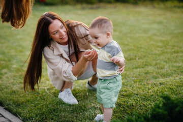 Fototapeta premium A young cute mother helps and teaches her little son to take his first steps during sunset in the park on the grass.