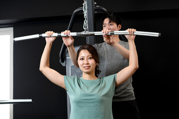 Naklejka premium ラットプルダウンをするアジア人女性と補助をする男性トレーナー
