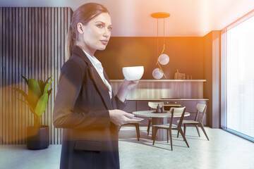 Attractive businesswoman in formal suit is drinking coffee at corporate office kitchen. Modern kitchen set and dining area as facilities for employees. Sun rays filling the room. Panoramic window