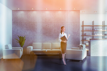 Attractive woman holding black folder with membership contract, waiting for clients in reception area of modern luxury spa wellness center to sign in documents. Towels, soaps and perfumes on shelves
