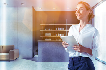 Smiling attractive woman holding tablet device and waiting for clients in reception area of modern luxury spa wellness center to sign in membership contract. Towels, soaps and perfumes are on shelves