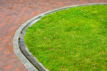 curved iron grid drainage system in the park by the green lawn meadow and walkway of red paving slabs with copy space, nobody.