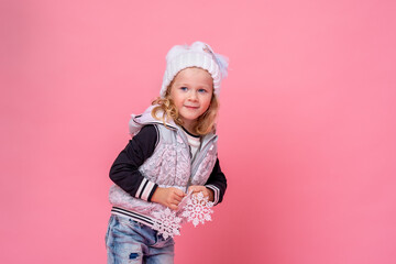 cute little girl in a hat and vest holds Christmas snowflakes on a pink background. the child is happy, having fun and getting ready for Christmas. The advertiser. A place to copy.