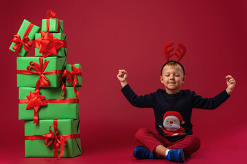 little boy in Santa Claus hat sits next to pyramid of Christmas gift boxes on red background in...