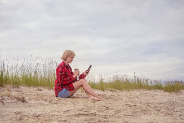 Naklejka na ściany i meble A young fair-haired Caucasian girl sits on the sand by the river and looks into a smartphone. The concept of outdoor recreation, relaxation.