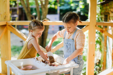 Happy sister kids play with sand and water in sensory baskets on the outdoor sensory table, sensory early development, montessori. Toddler and Big Sister Play with Sand, Soft Focus - obrazy, fototapety, plakaty