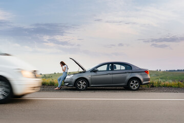 Fototapeta na wymiar A frustrated young girl stands near a broken-down car in the middle of the highway during sunset. Breakdown and repair of the car. Waiting for help.