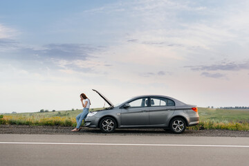 A frustrated young girl stands near a broken-down car in the middle of the highway during sunset. Breakdown and repair of the car. Waiting for help. - obrazy, fototapety, plakaty