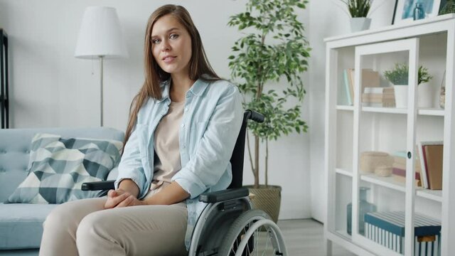 Portrait of young disabled woman sitting in wheelchair at home and looking at camera with light smile. Handicapped people and emotions concept.