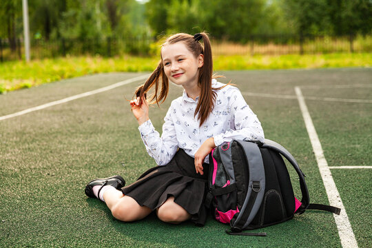 Smiling schoolgirl in a white blouse and black skirt sits on the green floor of the school stadium, long hair is taken in tails, briefcase, daytime, street, education