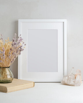 Empty white frame, glass vase with dried flowers, book, and natural crystal in a light interior. Mock up poster with place for your design.