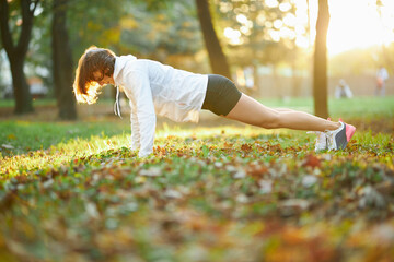 Fitness woman with dark hair wearing white sport jacket and black shorts standing in plank position...