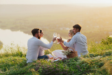 Friends have fun sitting on the bedspread on the hill at summer picnic. They are clinking glasses.