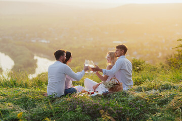 Group of young friends at summer sunset are sitting on the mountain and clinking glasses with wine. 