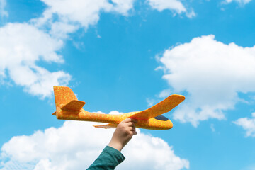 Hand of child holding a toy plane flying over the cloudy blue sky background. Adventure, travel and vacation concept.