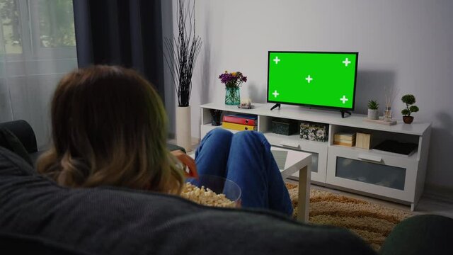 Woman Sitting on a Couch Home Watching Green Chroma Key Screen, Relaxing. Girl in a Cozy Room Watching Sports Match, News, Sitcom TV Show or a Movie on Green Screen eating popcorn.