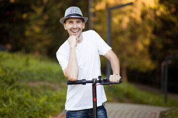 Portrait of young handsome man in white shirt, sunglasses and hat ready to ride his electric scooter bike on city street in sunny summer. Ecological transport concept. Electric urban transportation.