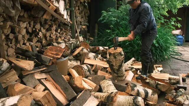 A man works in the courtyard of a cottage, next to a large wood-log, he cuts a log in half with a cleaver. A young man chops wood with an ax in the courtyard of a country house in the village.