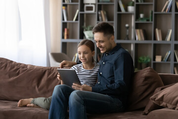 Happy caring young father sit rest on sofa in living room with teenage daughter use modern tablet...