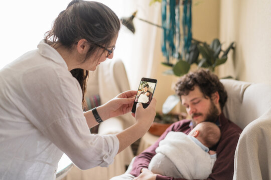 Smiling woman taking photo of husband with baby on smartphone