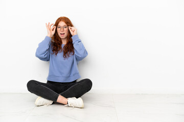 Fototapeta na wymiar Teenager redhead girl sitting on the floor isolated on white background with glasses and surprised
