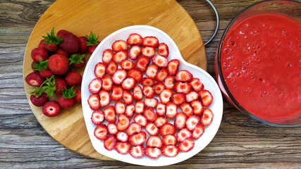 Strawberry slices on a white plate in the shape of a heart. Kluunichny puree in a glass dish. Whole red berries with leaves on a wooden background.