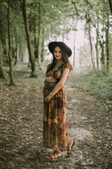 Beautiful young pregnant Caucasian woman, wearing long dress and black stylish hat, relaxing walking in the summer forest